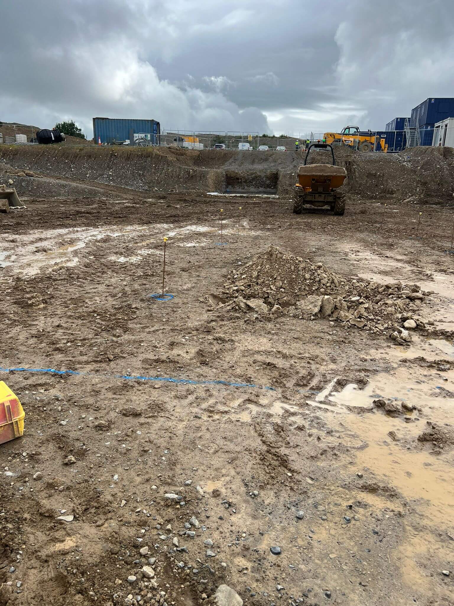 A muddy construction site with blue containers, yellow pipes, and a bulldozer
